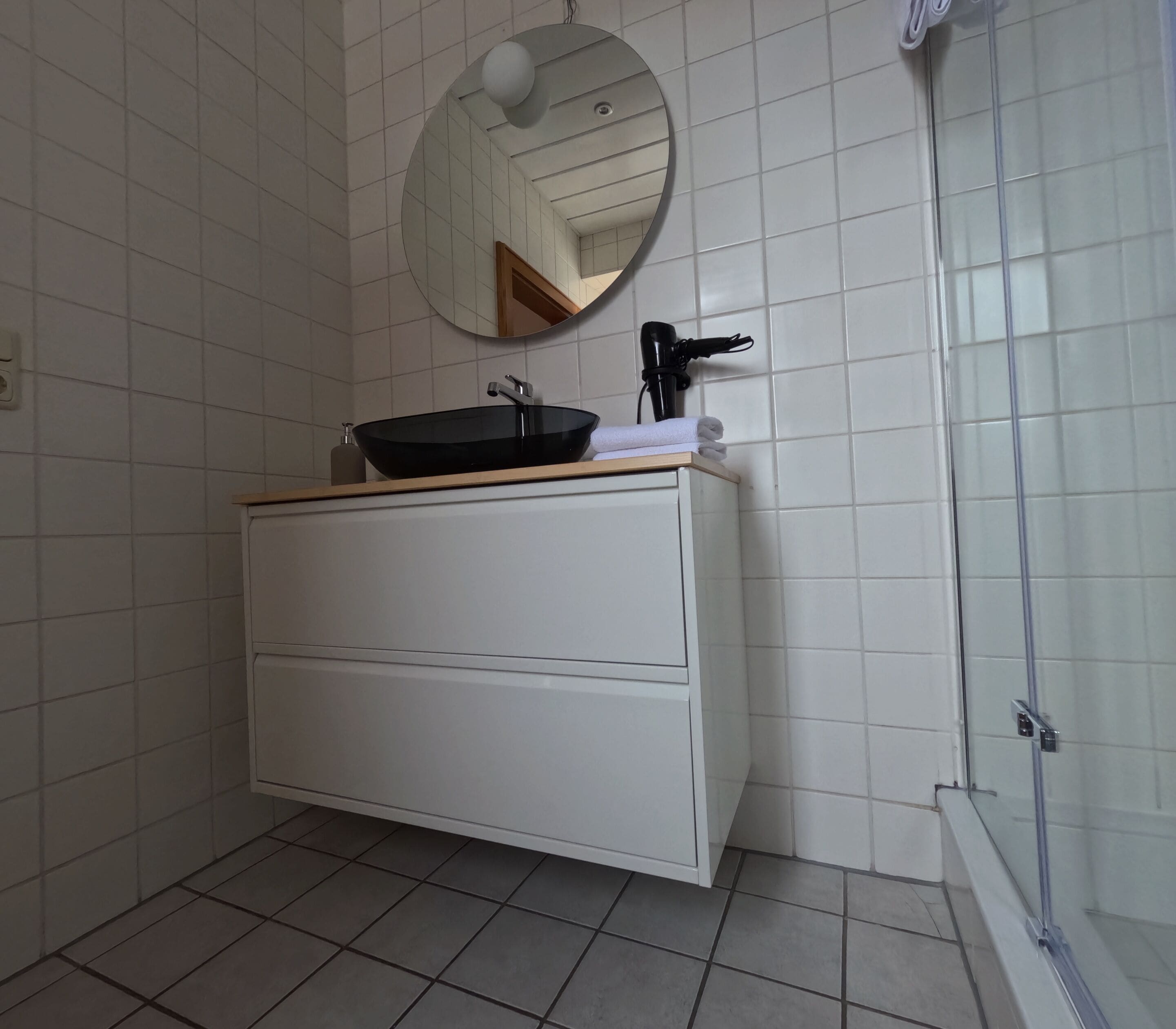 A bathroom with a white vanity, a black sink, a round mirror, and a shower area.