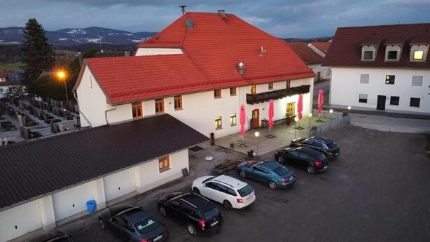 A building with a red roof and white walls, a parking area with several cars, and a view of mountains in the background. - Gasthof & Pension Zum Zacherwirt (Hengersberg)