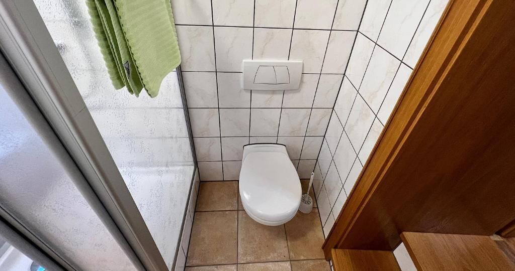 A small bathroom with a white toilet, a green towel hanging on the wall, and a light switch above the toilet.