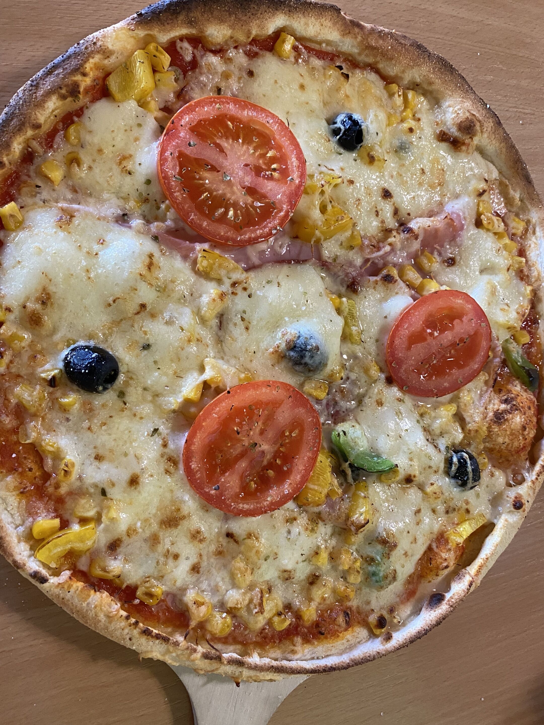 A pizza with tomatoes, olives, and corn on a wooden surface.