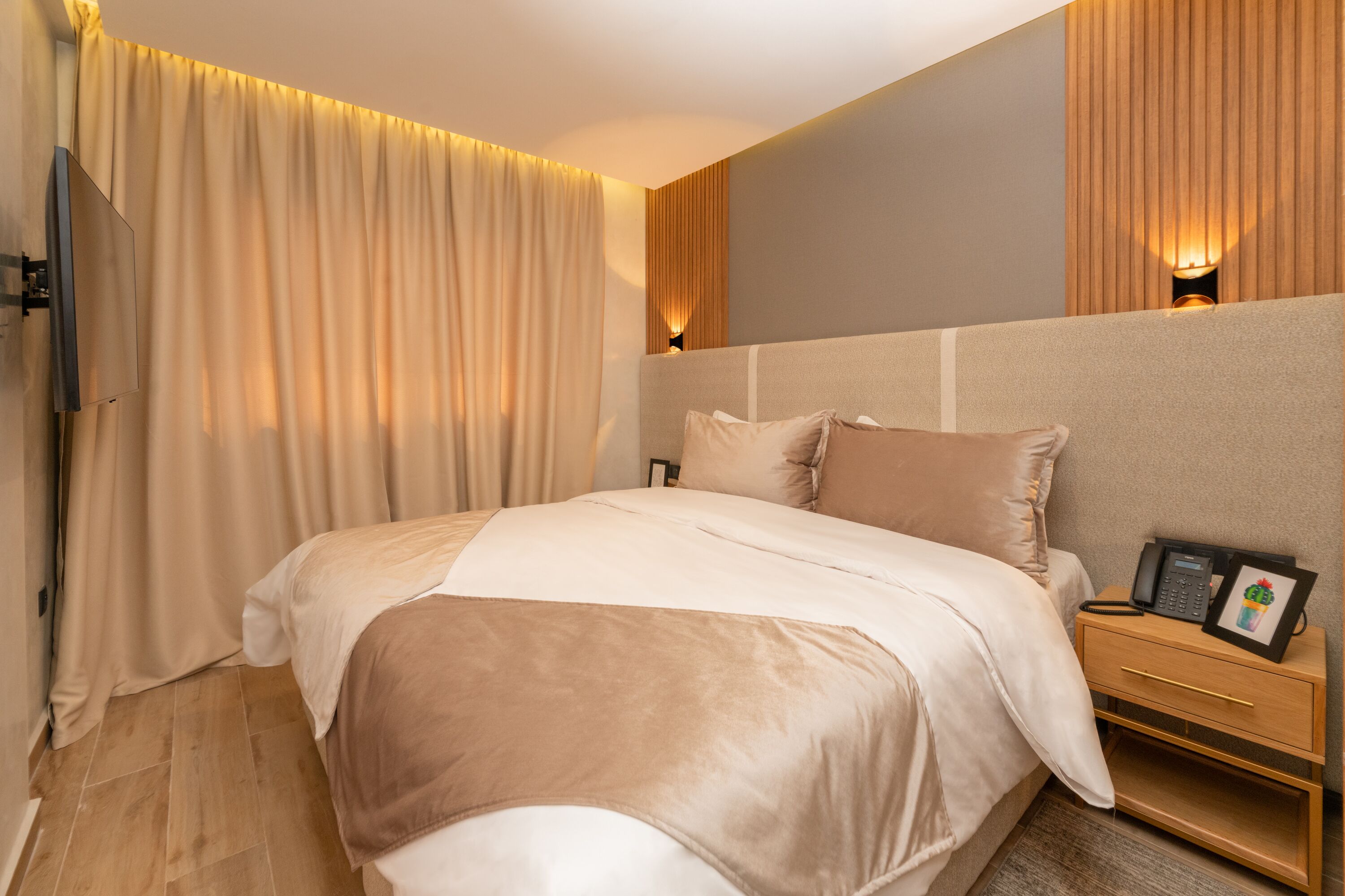 A modern hotel room with a large bed, a bedside table with a phone and picture frame, and a wall-mounted TV.