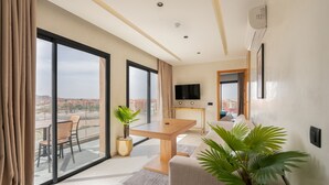 Quarto de hotel moderno com uma grande porta de vidro deslizante que dá para uma varanda, uma mesa de jantar de madeira, um sofá e uma televisão de tela plana.
