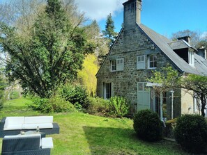 A stone house with a sloped roof, a chimney, and a garden with a sofa and table.
