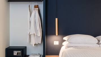 Un dormitorio con cama, mesita de noche, ropa colgada y una lámpara fijada a la pared.