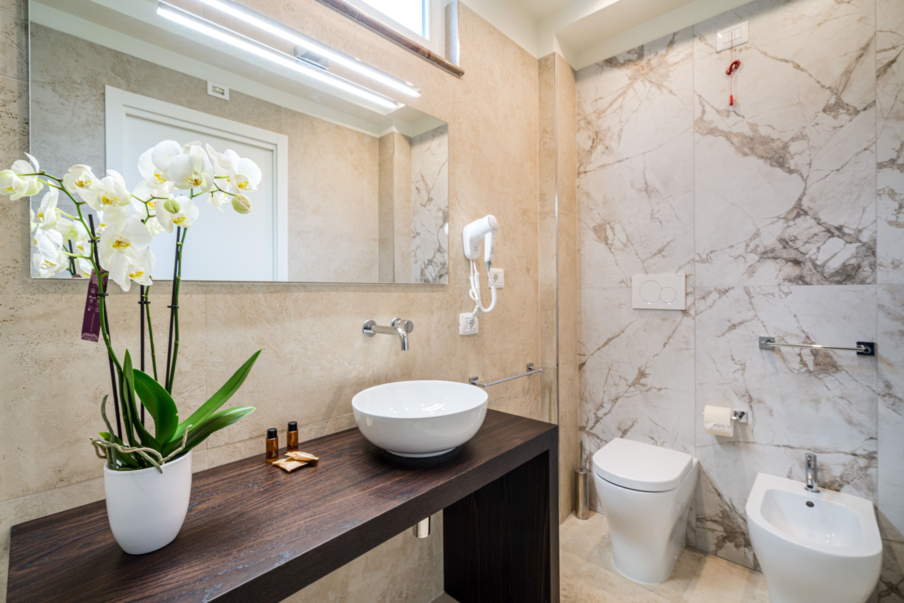 Une salle de bain moderne avec un mur en marbre, un lavabo blanc, un bidet et un sèche-cheveux fixé au mur.