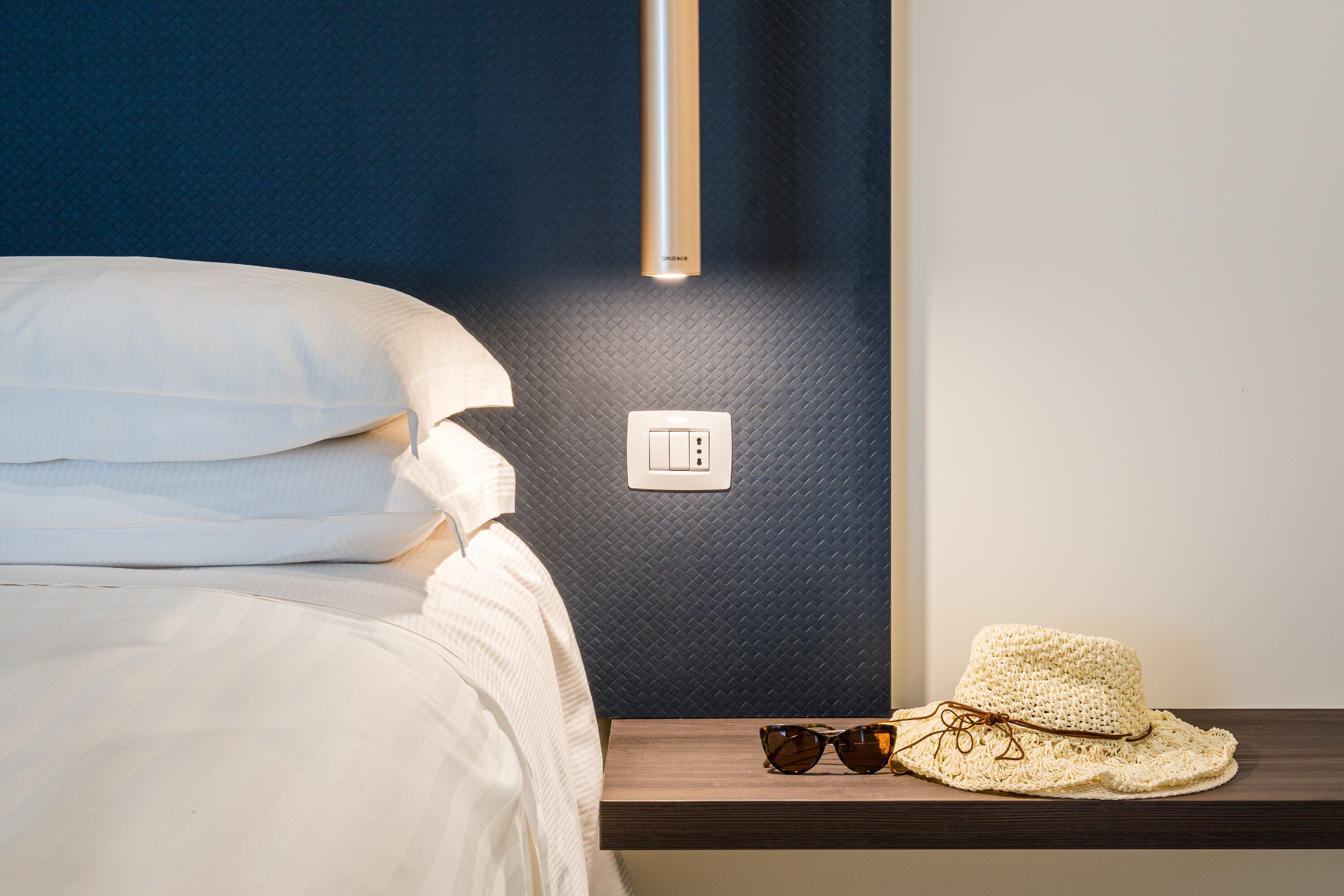 Una cama bien hecha con ropa blanca, una mesita de noche con un sombrero y gafas de sol, y un interruptor de pared.