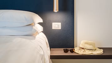 A neatly made bed with white linens, a bedside table with a hat and sunglasses, and a wall switch.