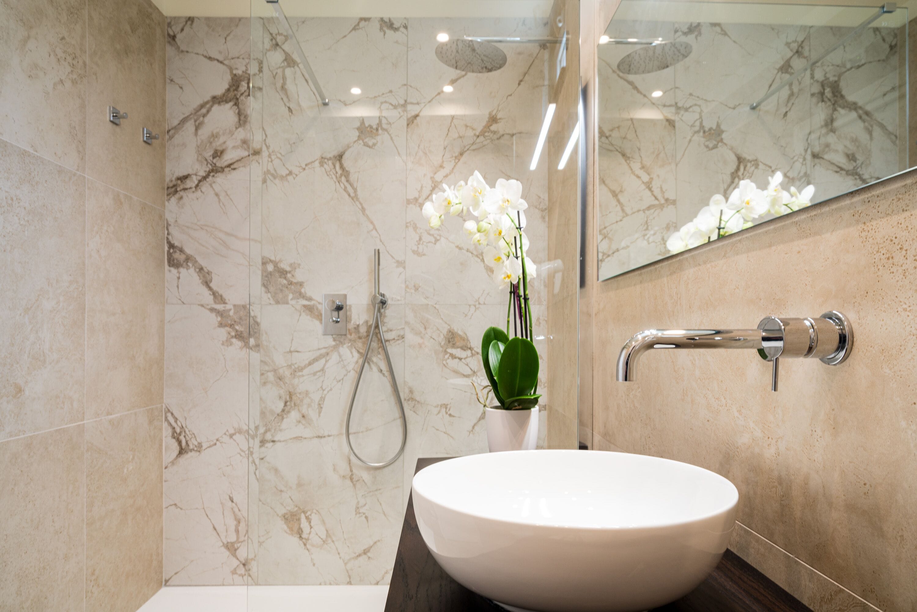 Un baño con pared de mármol, lavamanos blanco, planta verde y ducha.