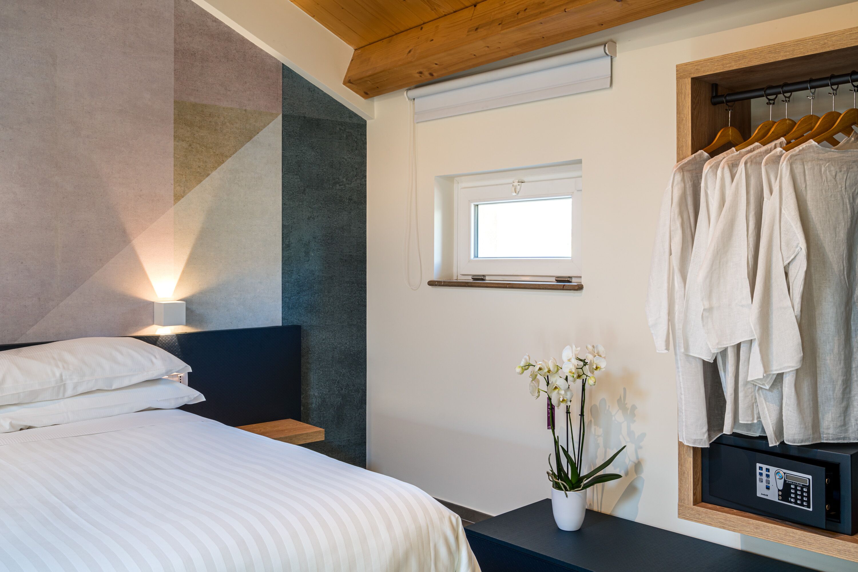A bedroom with a bed, a bedside table with a plant, a window, and a clothing rack.