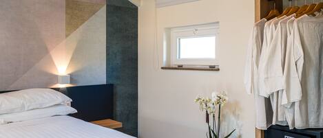 A bedroom with a bed, a bedside table with a plant, a window, and a clothing rack.