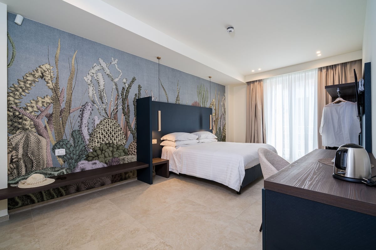 Een moderne hotelkamer met een grote muurschildering van zeedieren, een bed met donker hoofdeinde, een nachtkastje en een uitzicht op de stad door grote ramen.