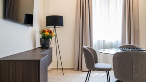 A modern room with a dark wood desk, a glass table, two grey chairs, a floor lamp, and a large window with curtains.