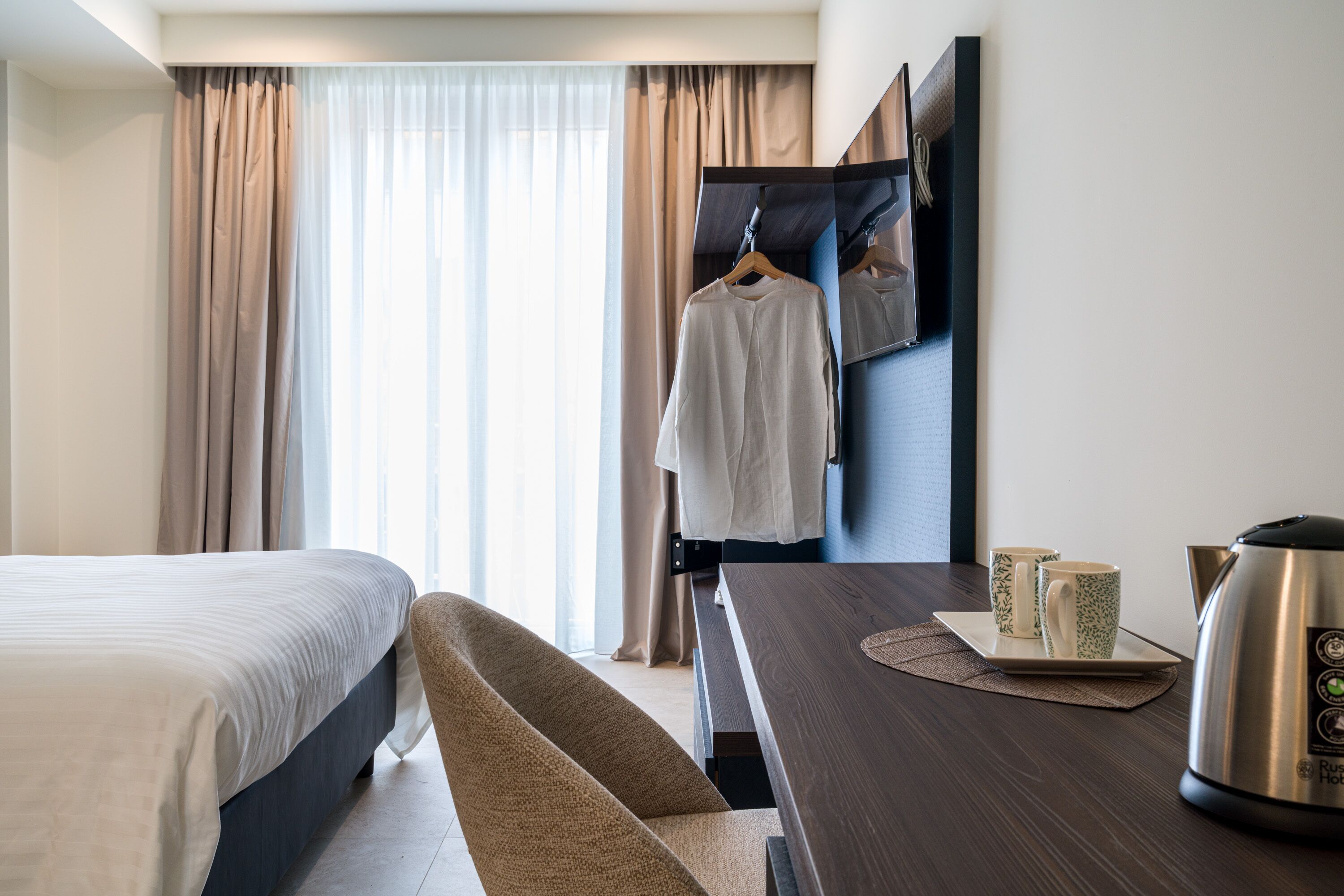 Ein Hotelzimmer mit einem Bett, einem Holztisch mit Wasserkocher und zwei Tassen, einem Sessel und einem Kleiderschrank mit Spiegel.