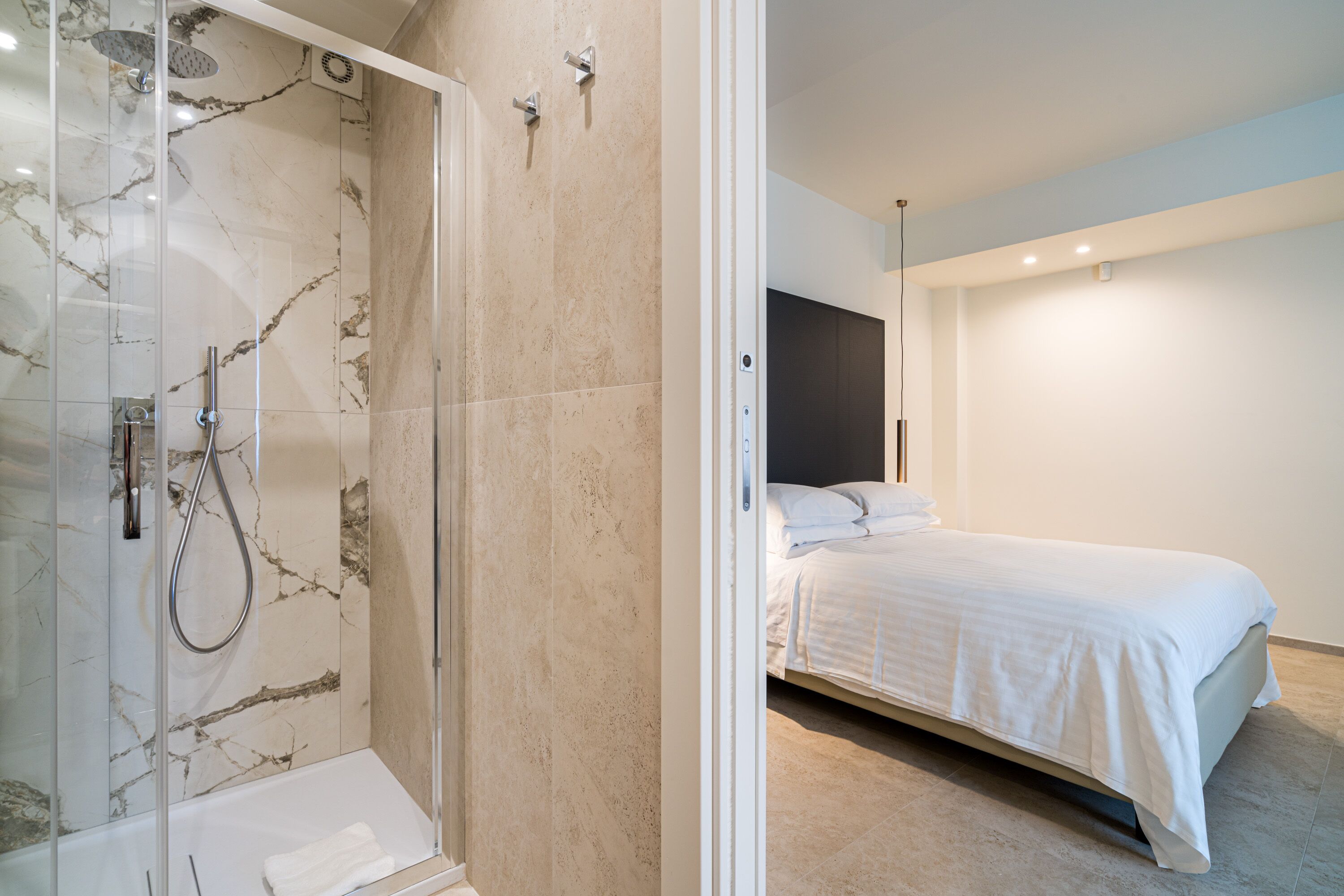 A modern bathroom with a glass-enclosed shower and a separate bedroom with a bed and a nightstand.