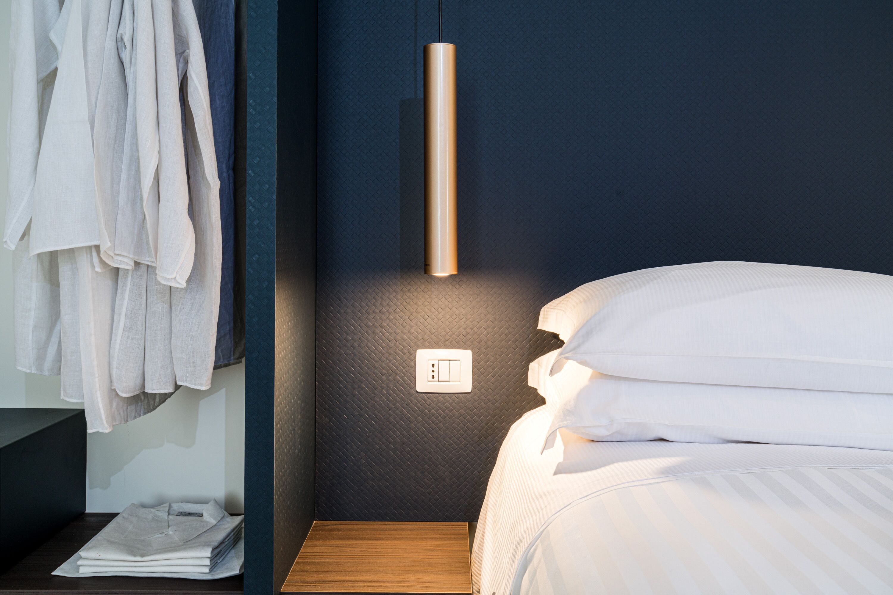 A neatly made bed with white linens, a hanging light fixture, and a wall socket.