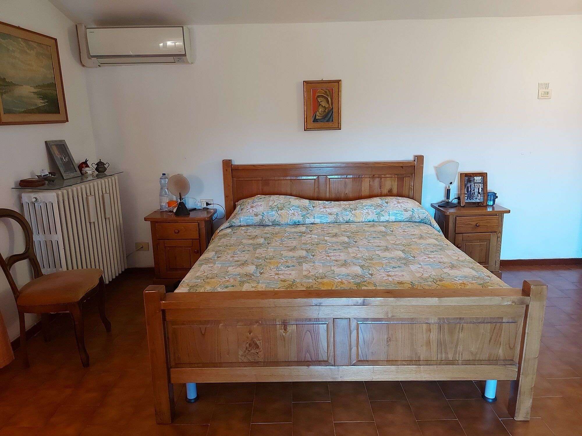 A bedroom with a wooden bed, a nightstand with a lamp, a chair, a radiator, and a wall-mounted air conditioner.