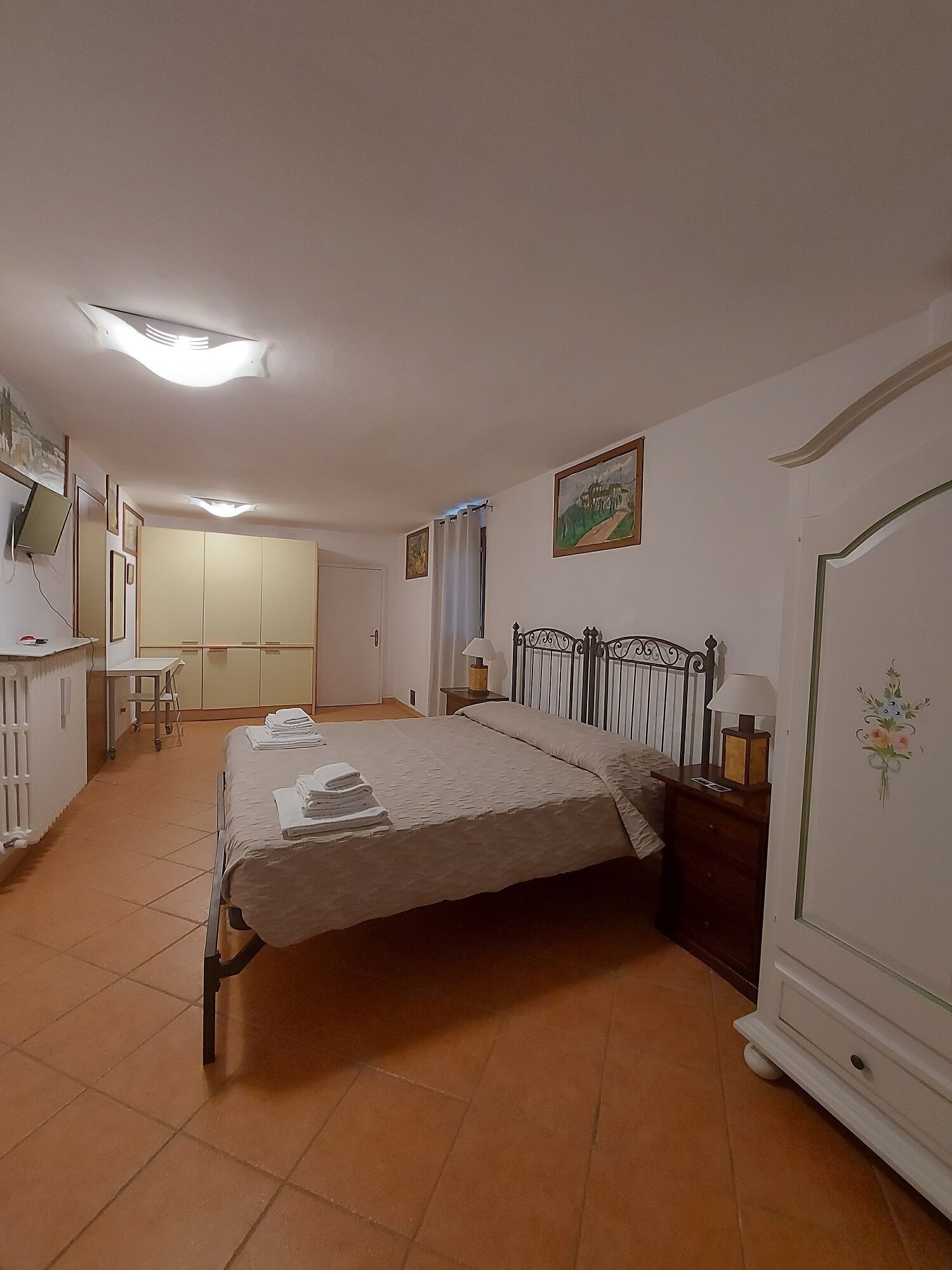 A bedroom with a bed, bedside table, lamp, and wardrobe. There is a desk with a chair and a TV mounted on the wall.