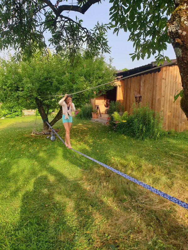 En person går over et blåt reb i en græsplæne med træer og en træbygning i baggrunden.