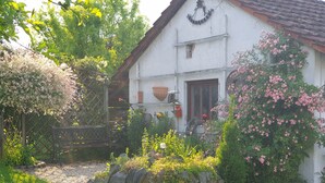 A white building with a sloped roof, surrounded by a garden with various plants and flowers. - Appartement mit gr. Garten u. 2 Bäder (Vilshofen an der Donau)