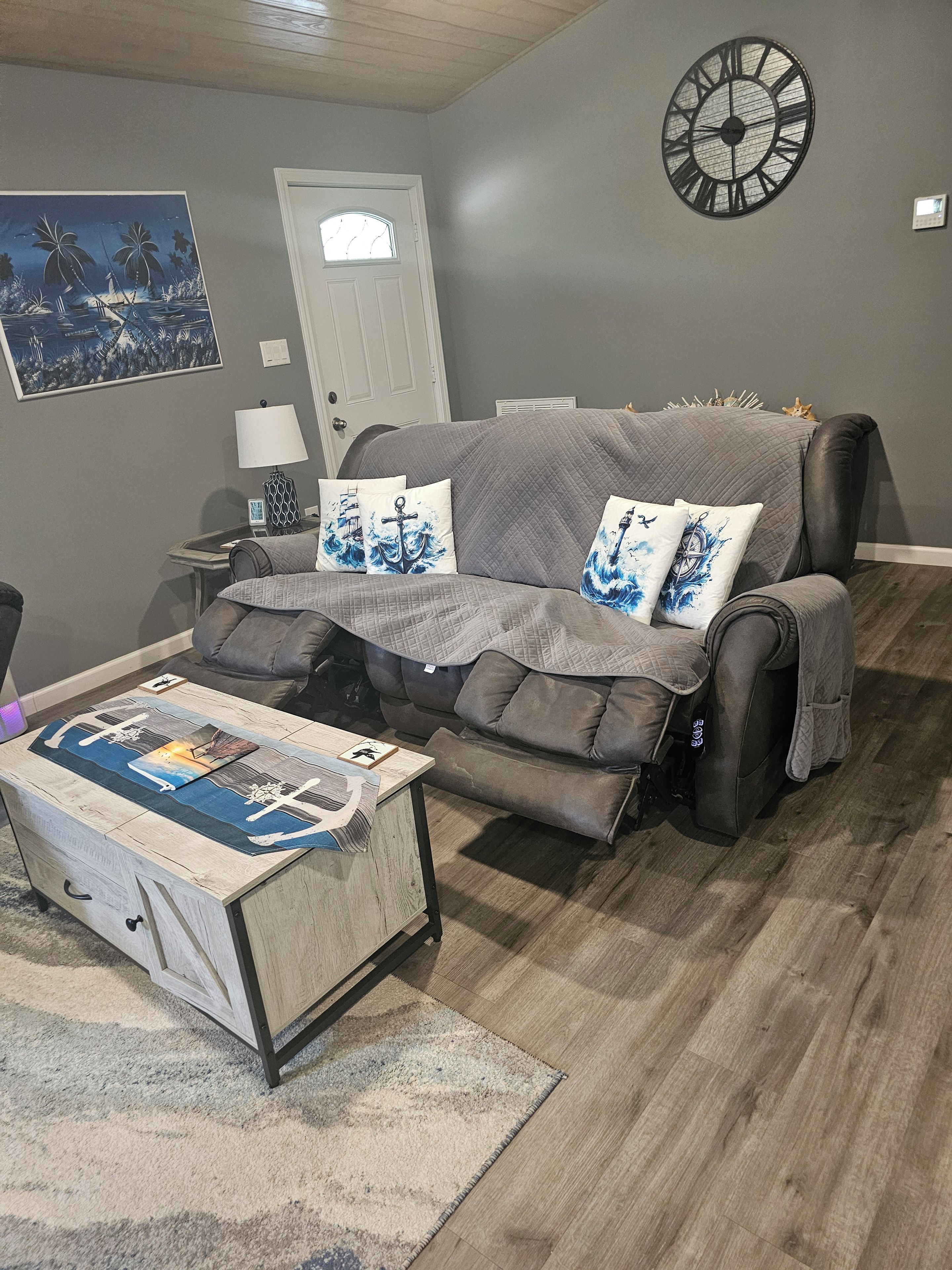 A living room with a grey sofa, a coffee table with a nautical-themed rug, a wall clock, and a door with a small window.