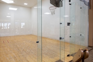 Una cancha de squash con paredes de vidrio, piso de madera y aro de baloncesto.