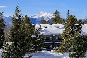 Edificio cubierto de nieve con un balcón, rodeado de árboles de hoja perenne y una cadena montañosa al fondo.
