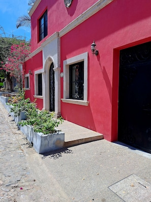 A red building with a white door and windows, a black gate, and potted plants along the sidewalk.