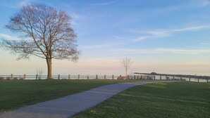 Een geplaveid pad in een park met een grote boom, een hek en een verre blik op een waterpartij.