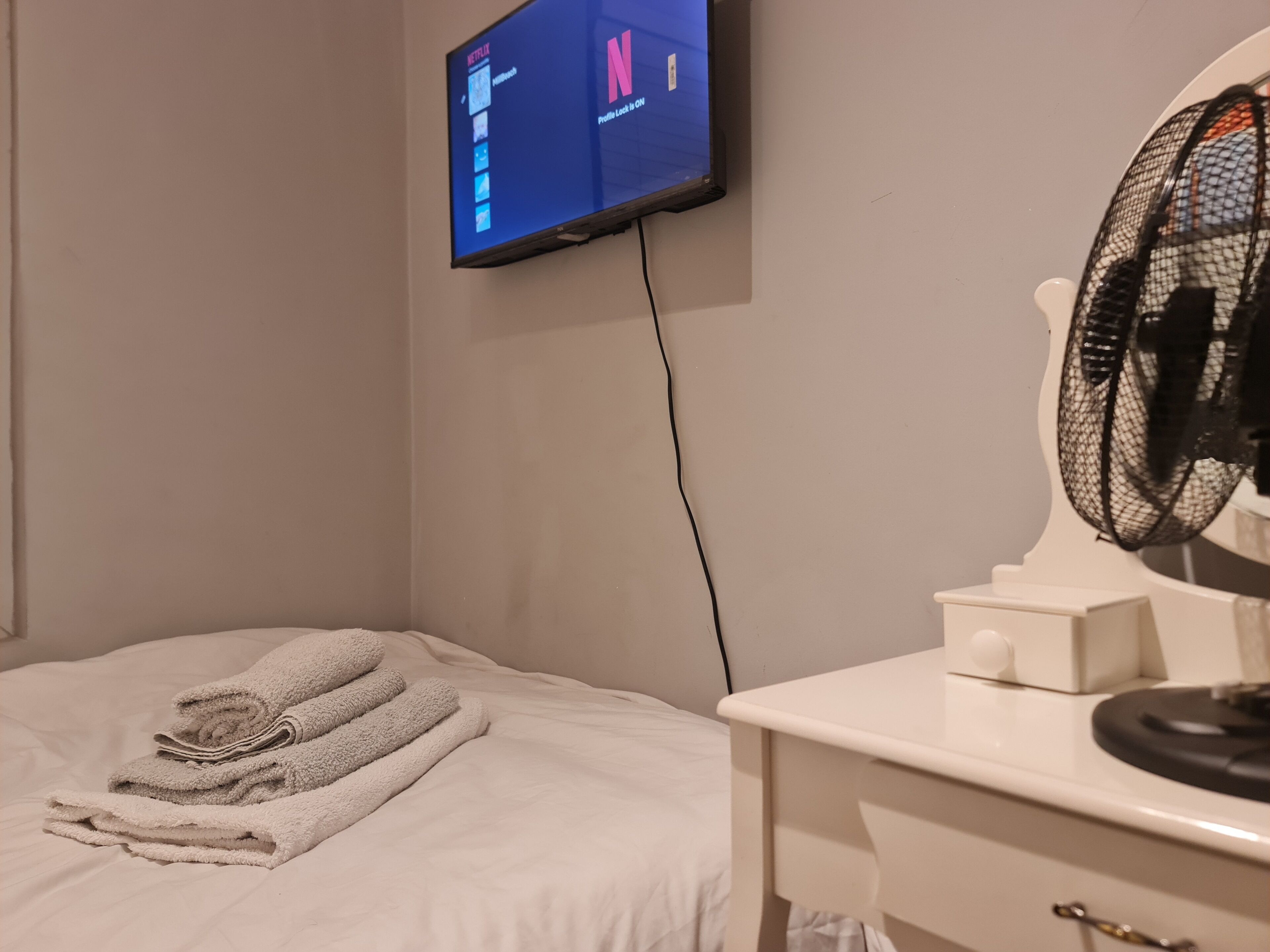 Ett hotellrum med en säng, en tv som är monterad på väggen och visar Netflix, ett vitt nattduksbord med en fläkt och prydligt hopvikta handdukar.