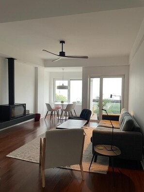 A modern living room with a fireplace, a ceiling fan, a sofa, a coffee table, and a dining area with chairs and a table. - Oceanview 4 U - Close to Lisbon! (Oeiras)