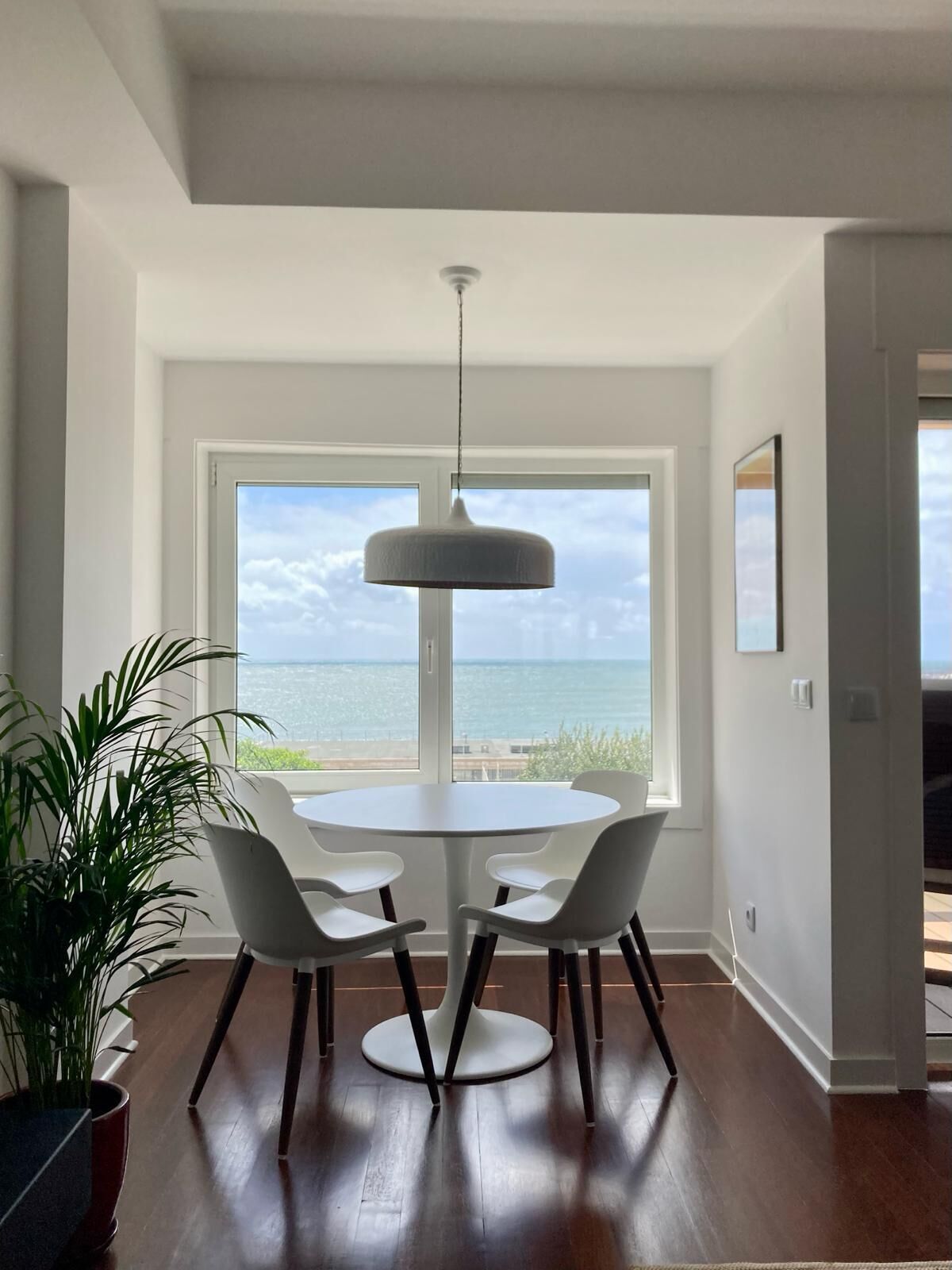 Uma área de refeições com uma mesa redonda e quatro cadeiras, um lustre pendurado e vista para o oceano através de grandes janelas.
