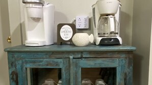 Un mobiletti color petrolio con due ripiani, ognuno contenente tazze bianche e una macchina per il caffè. Sopra il mobiletti è appesa una cornice con un'immagine.