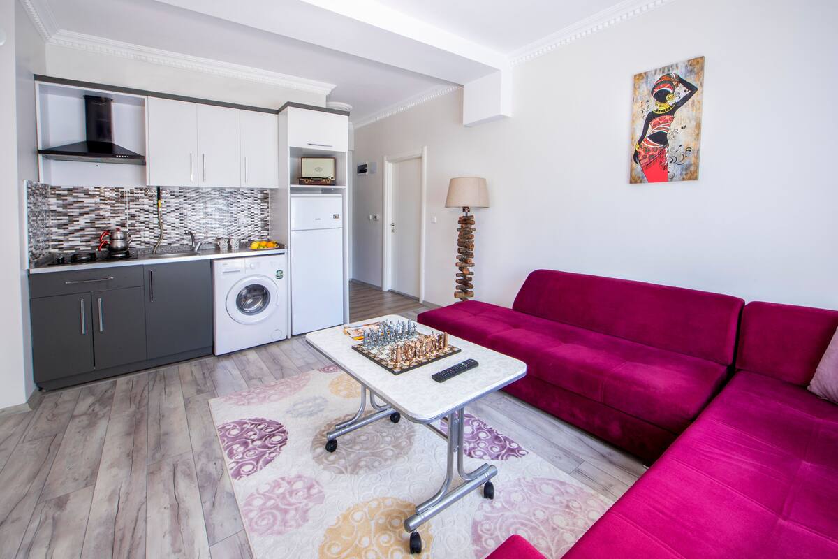 A modern apartment with a kitchen, a living area with a red sofa, and a chessboard on a mobile table.