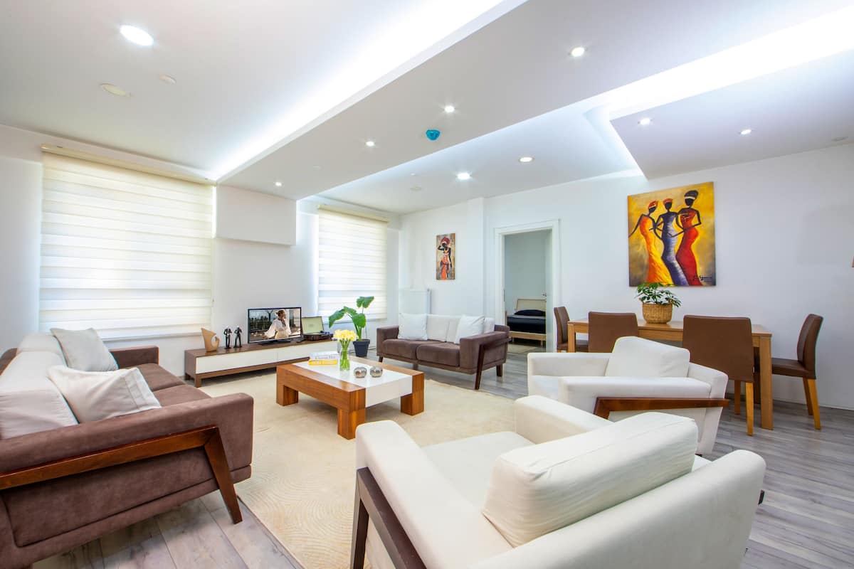 A modern living room with a brown sofa, white armchairs, a wooden coffee table, and a dining area with a table and chairs.