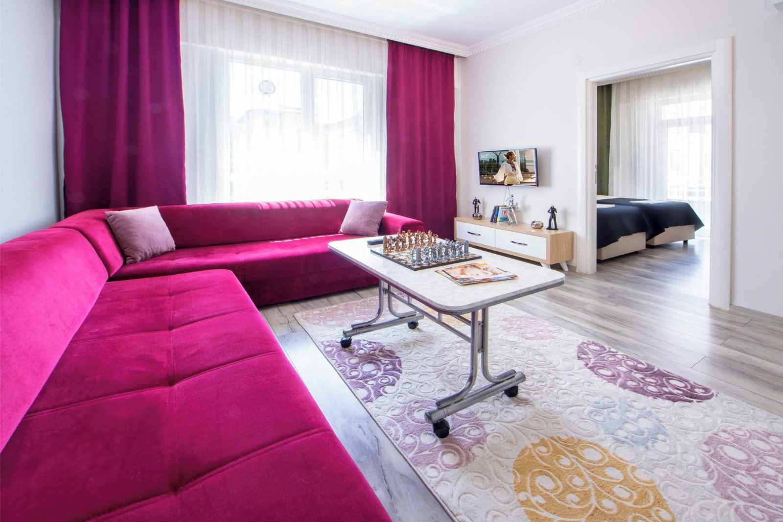 A modern living room with a pink sofa, a coffee table with chess pieces, and a bed in the background.