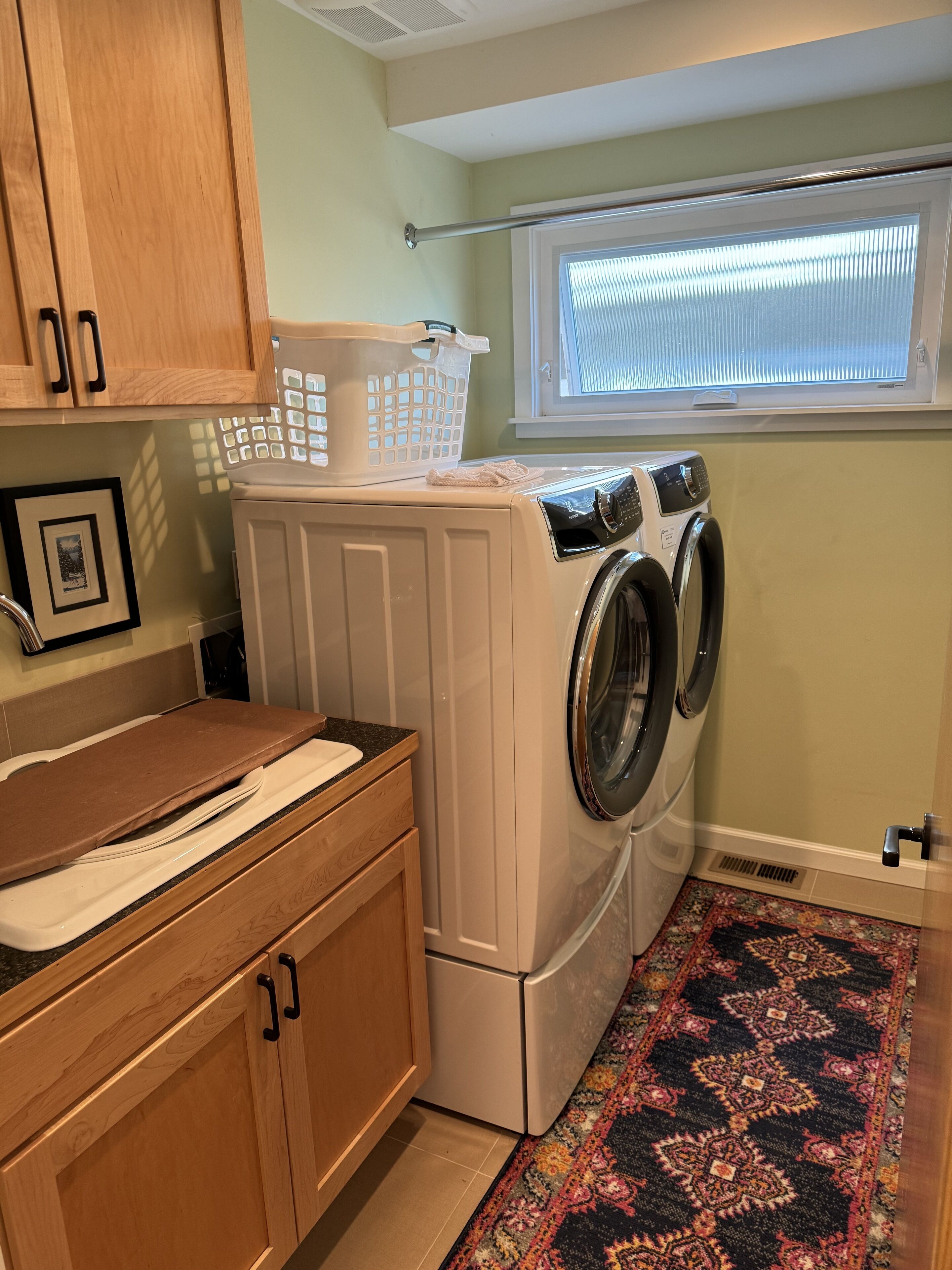 Un espace buanderie comprenant un lave-linge et un sèche-linge à chargement frontal, des armoires en bois, un plan de travail, une fenêtre avec des stores et un tapis à motifs.