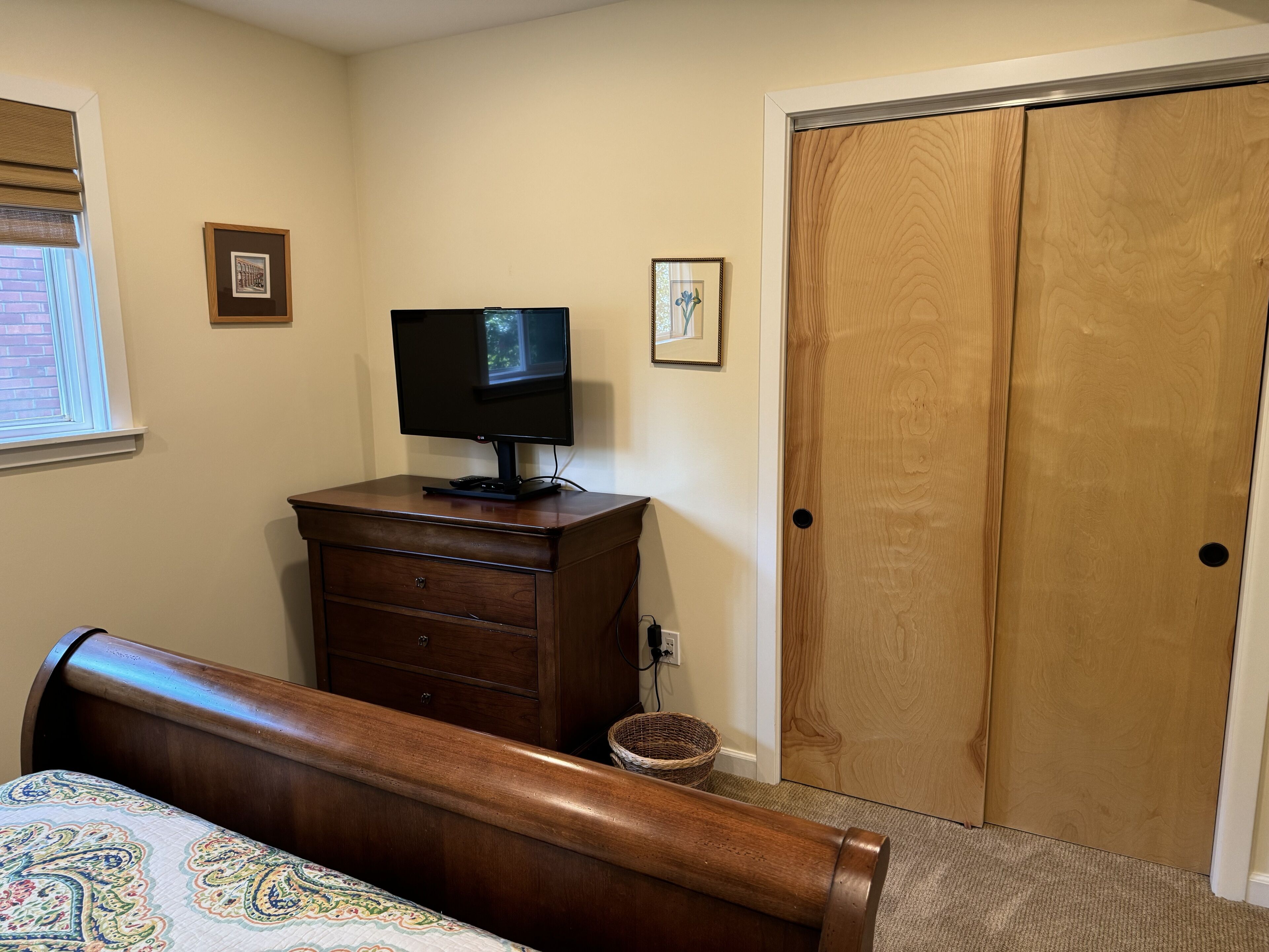 A bedroom with a wooden dresser, a television on top, a bed with a patterned bedspread, a wooden door, and a window with blinds.