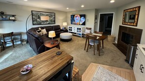 A living room with a sofa, ottoman, coffee table, TV, fireplace, and a dining area with chairs and a table. - Enjoy all the comforts of home in a safe, wooded area of NE Seattle, close to UW (Seattle)