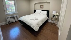 A neatly made bed in a room with a radiator, a window with a view of trees, and a small table with a lamp and flowers. - Cozy House,  cottage style in midtown Toronto. Great for Family. Pet friendly  (Toronto)