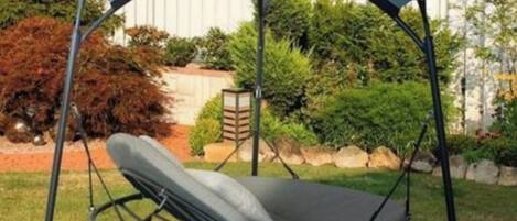 A corner swing chair with a canopy, set in a garden with a white fence and greenery in the background.