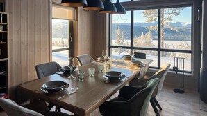 Ruokailutilassa on puinen pöytä, johon on katettu neljälle, suuret ikkunat, joista avautuu lumipeitteinen maisema, ja pöydän yläpuolella on riippuvalaisimet.