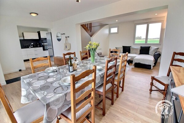 A dining area with a table set for six, surrounded by wooden chairs. A kitchen is visible in the background, and a bed is seen in the adjacent room.