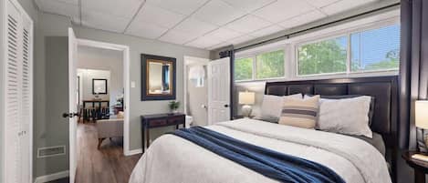 Un dormitorio con una cama grande, una mesita de noche, una ventana con cortinas y una puerta que da a otra habitación.