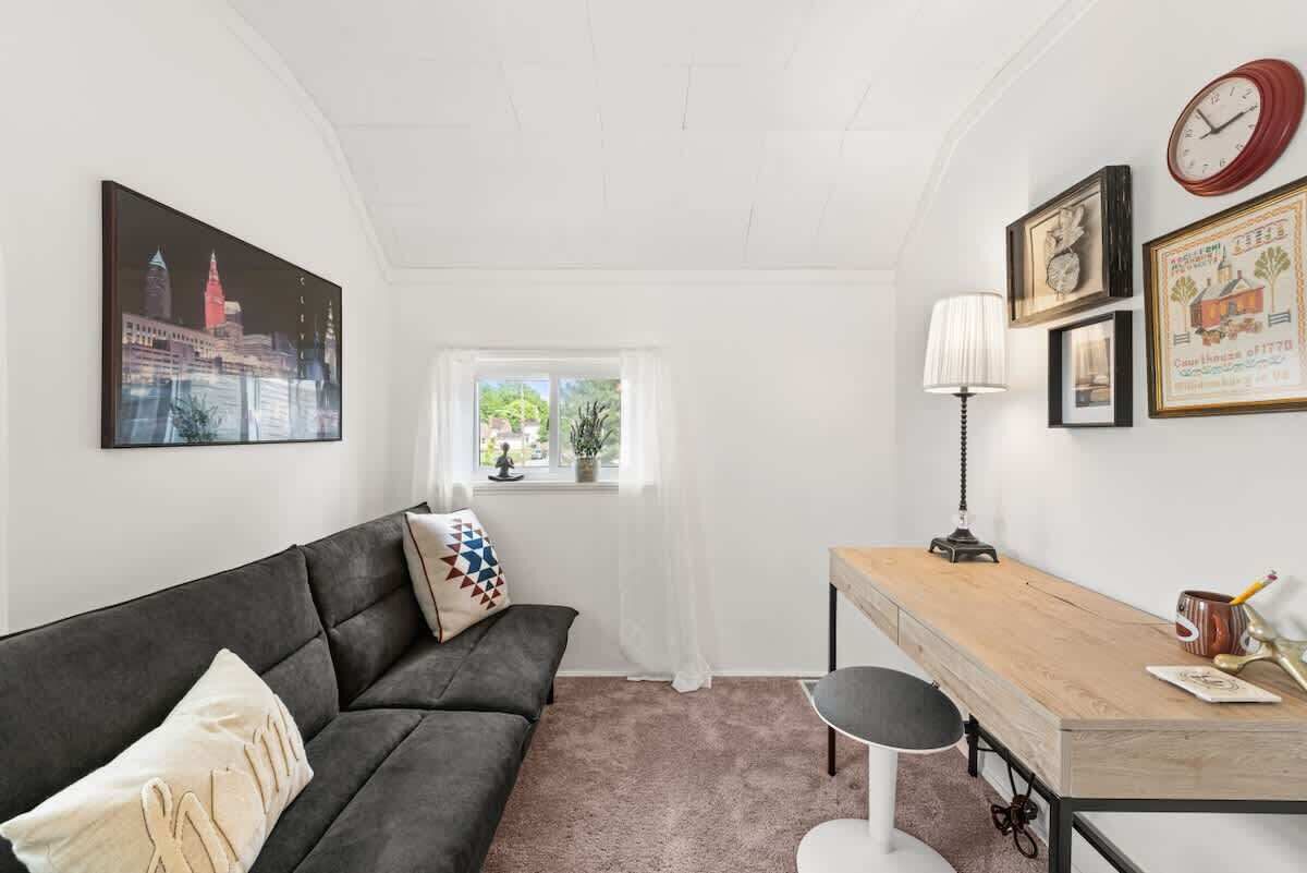 A modern living room with a dark grey sofa, a wooden desk, and a small window with sheer curtains.