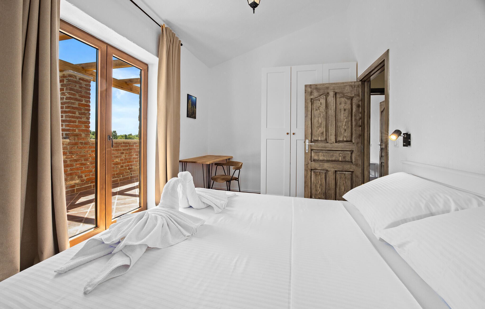 Une chambre avec un lit, une porte en bois, une petite table avec des chaises et une vue sur un mur de briques à travers une porte en verre.