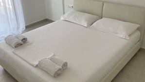 Ein ordentlich bezogenes Bett mit weißen Leinen und aufgerollten Handtüchern, ein schlichter Kopfteil und ein Fenster mit durchscheinenden Vorhängen.