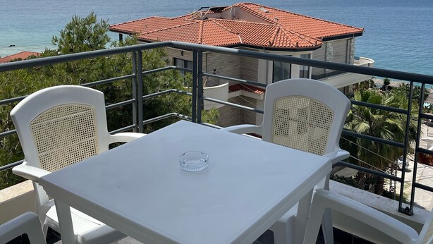 Ein Balkon mit weißem Tisch und Stühlen, mit Blick auf die Küste, im Hintergrund ein Gebäude mit rotem Dach.
