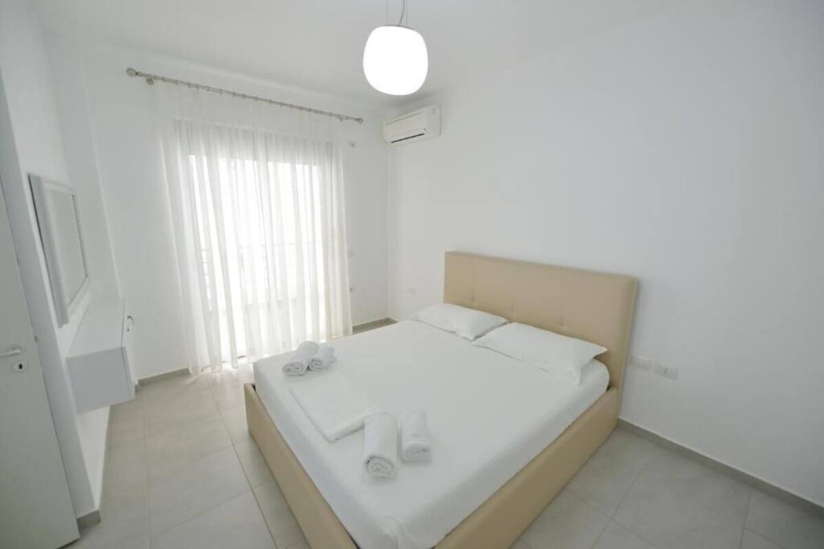 A clean, modern hotel room with a single bed, white linens, a flat-screen TV, and a window with sheer curtains.