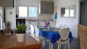Une cuisine de taille modeste, avec des meubles blancs, une table à manger pour quatre personnes recouverte d’une nappe à pois bleus, et une plante en pot posée sur le comptoir.