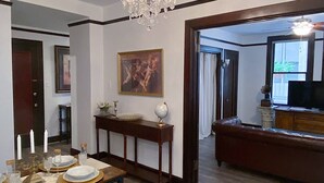Zona de menjador amb taula de fusta preparada per a quatre persones, un lampadari i vista a una altra habitació amb televisió.